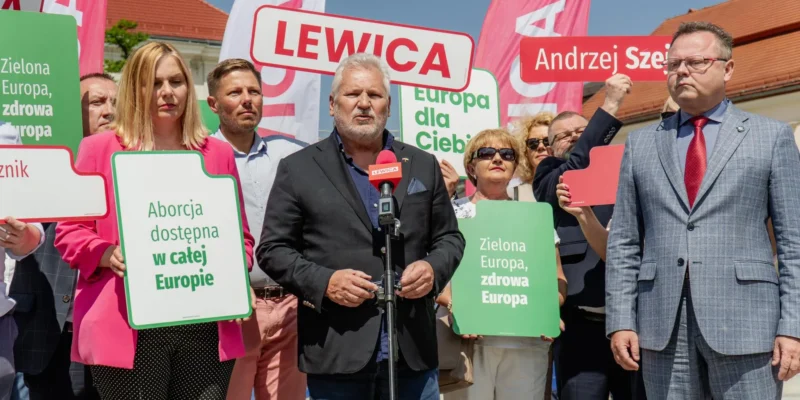 Grupa zwolenników Lewicy trzyma plakaty z hasłami „Zielona Europa, zdrowa Europa” i „Aborcja dostępna w całej Europie” podczas przemowy przy mikrofonie.