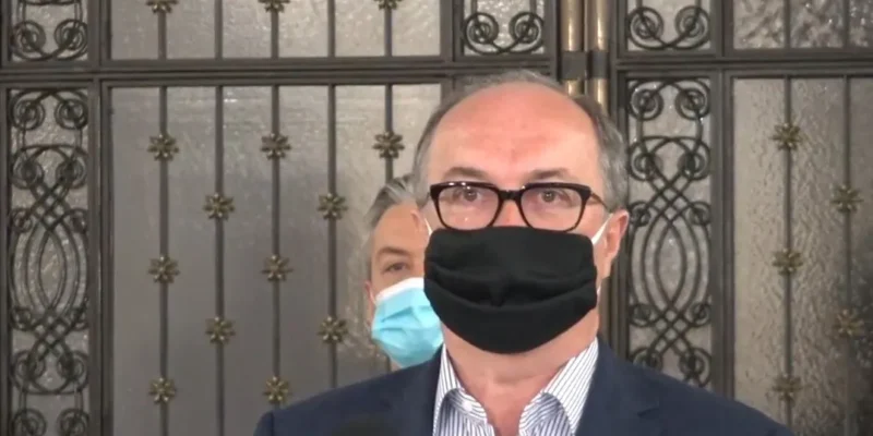 Polityk w okularach, czarnej maseczce i garniturze stoi przy mikrofonie podczas konferencji prasowej, na tle ozdobnej bramy.