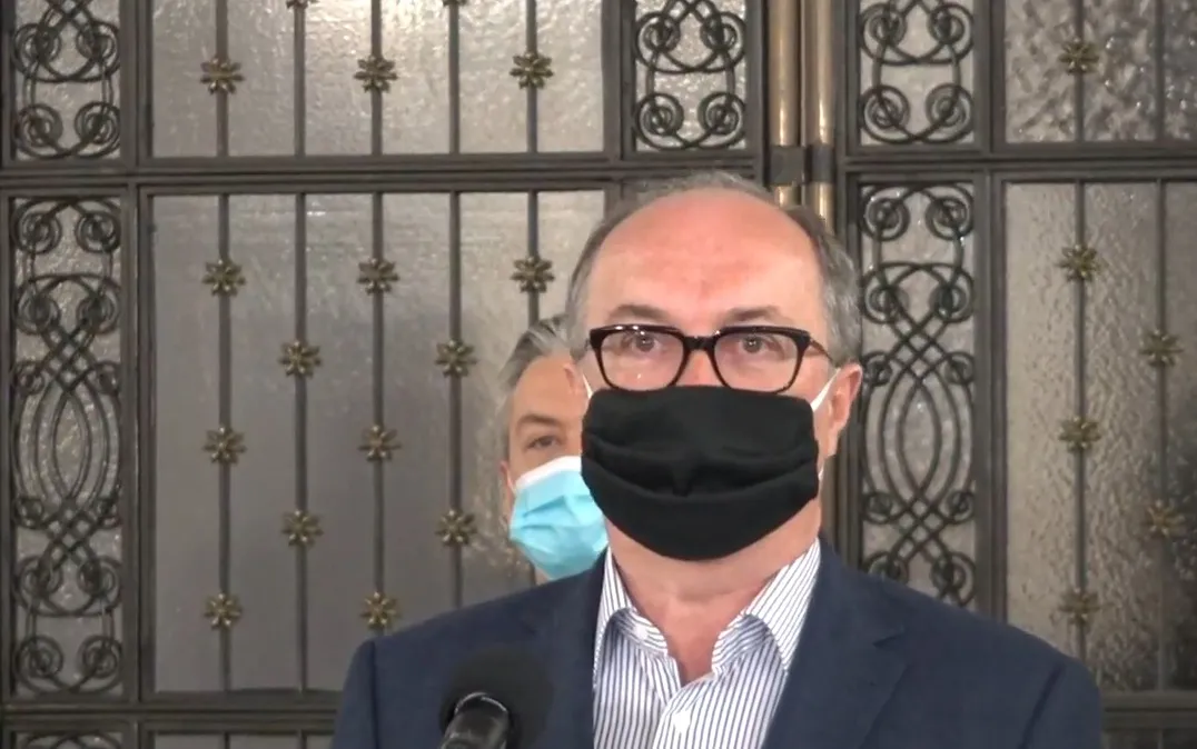 Polityk w okularach, czarnej maseczce i garniturze stoi przy mikrofonie podczas konferencji prasowej, na tle ozdobnej bramy.