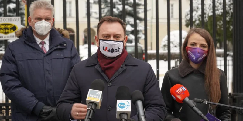 Trzy osoby w maseczkach stoją przed metalowym ogrodzeniem i przemawiają do mediów podczas konferencji prasowej na tle zimowego otoczenia.