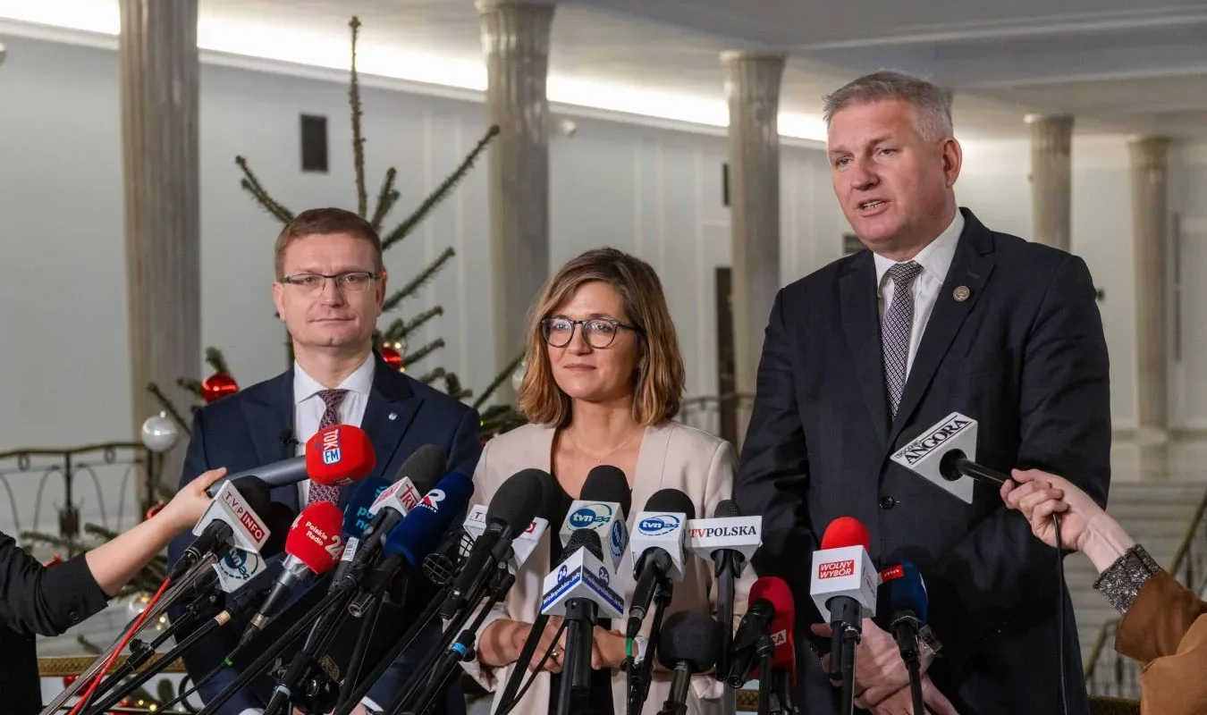 Trzy osoby w garniturach stoją przed licznymi mikrofonami podczas konferencji prasowej o refundacji in vitro z budżetu państwa, z choinką w tle.