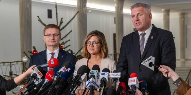 Trzy osoby w garniturach stoją przed licznymi mikrofonami podczas konferencji prasowej o refundacji in vitro z budżetu państwa, z choinką w tle.