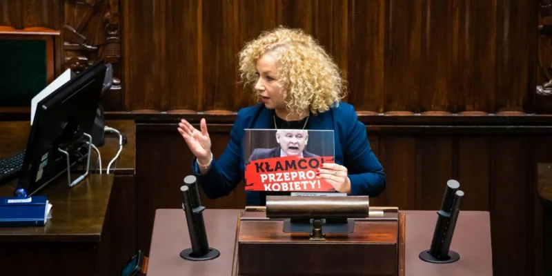 Kobieta w stylowym, niebieskim żakiecie trzyma plakat z napisem "Kłamco! Przeproś kobiety!" podczas wystąpienia w parlamencie.