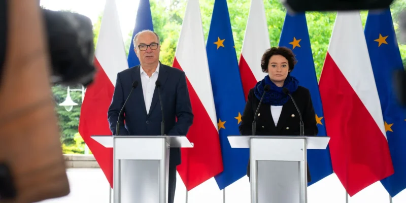 Dwóch polityków stoi przy oddzielnych podiumach z mikrofonami podczas konferencji prasowej, na tle flag Polski i Unii Europejskiej.