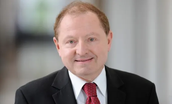 Portret mężczyzny w średnim wieku w ciemnym garniturze, białej koszuli i czerwonym krawacie, uśmiechniętego na jasnym, rozmytym tle.