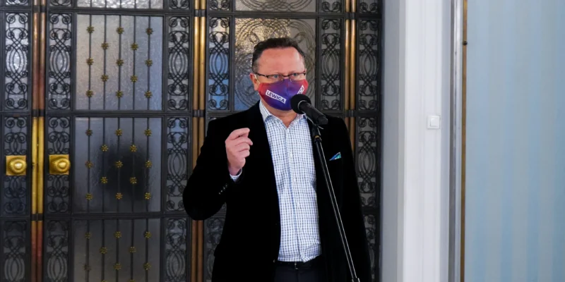 Mężczyzna w czarnej marynarce i koszuli stoi przy mikrofonie, nosi kolorową maseczkę z napisem LEWICA i stoi przed ozdobną metalową bramą.