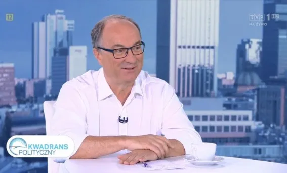 Uśmiechnięty mężczyzna w białej koszuli i okularach siedzi przy stoliku podczas programu politycznego, z filiżanką i dokumentami na tle miejskiej panoramy.