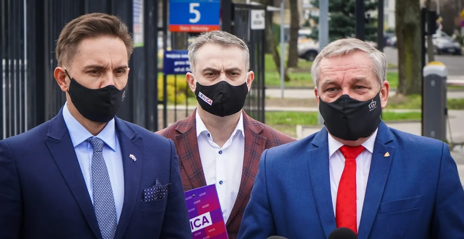 Trzech mężczyzn w garniturach i maseczkach stoi przed kamerami podczas konferencji prasowej.