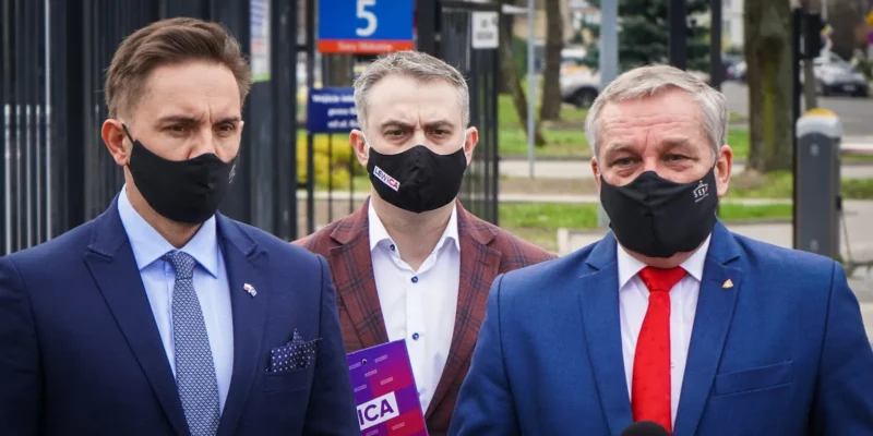 Trzech mężczyzn w garniturach i maseczkach stoi przed kamerami podczas konferencji prasowej.