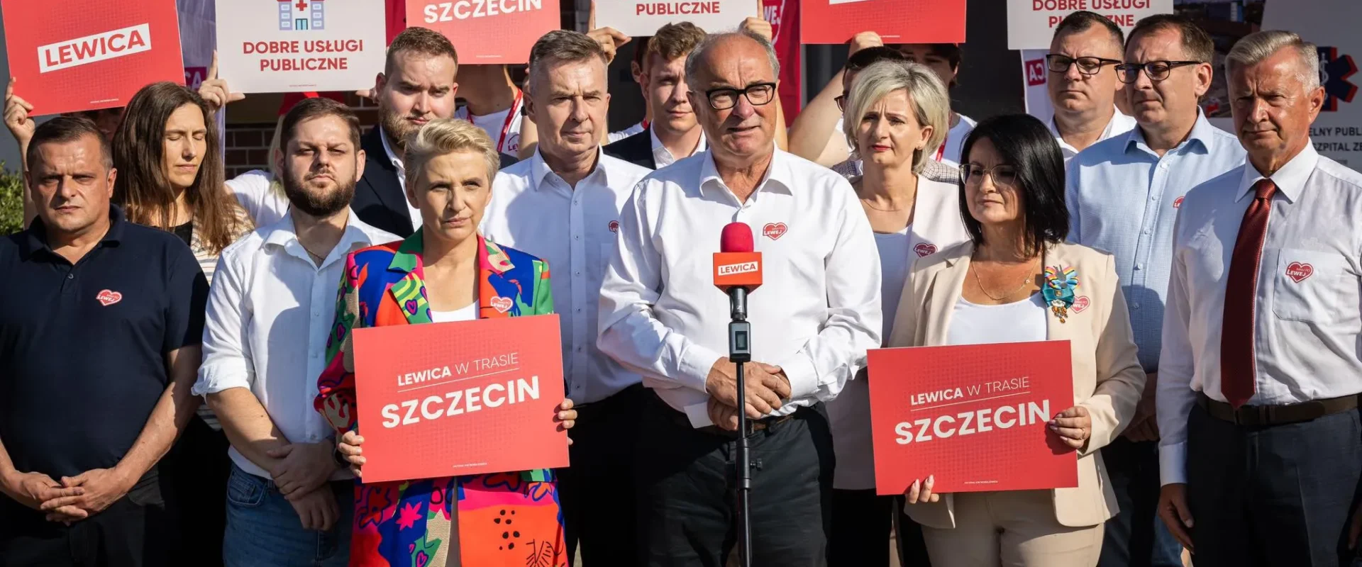 Grupa polityków Lewicy podczas konferencji prasowej, stojąca z czerwonymi plakatami z hasłami "Lewica" i "Dobre usługi publiczne" przy mikrofonie z logiem Lewicy.
