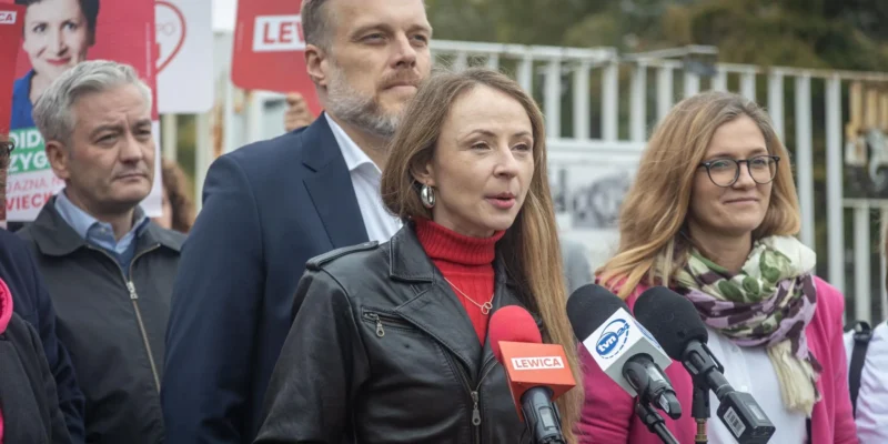 Kobieta w czerwonym golfie i czarnej skórzanej kurtce przemawia do mikrofonów podczas konferencji prasowej, w tle widoczni są inni działacze i banery Lewicy.