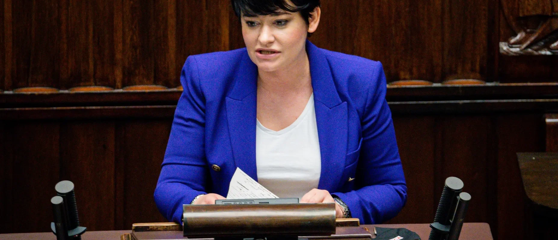 Kobieta z krótkimi czarnymi włosami w niebieskiej marynarce siedzi przy biurku w sali posiedzeń i trzyma kartki.