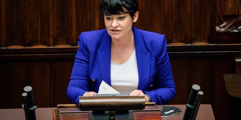 Kobieta z krótkimi czarnymi włosami w niebieskiej marynarce siedzi przy biurku w sali posiedzeń i trzyma kartki.