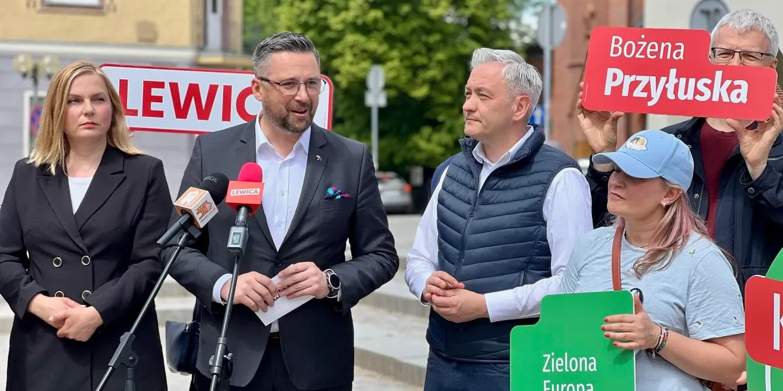 Grupa osób na zewnątrz prowadzi konferencję prasową; mężczyzna w garniturze przemawia do mikrofonów, obok niego stoi kobieta w czarnym garniturze, a po prawej widoczni są zwolennicy trzymający plakaty z hasłami wyborczymi.