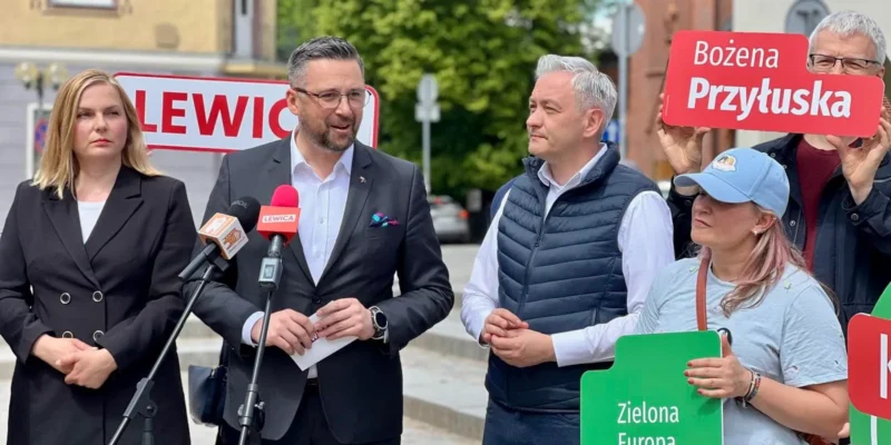 Grupa osób na zewnątrz prowadzi konferencję prasową; mężczyzna w garniturze przemawia do mikrofonów, obok niego stoi kobieta w czarnym garniturze, a po prawej widoczni są zwolennicy trzymający plakaty z hasłami wyborczymi.