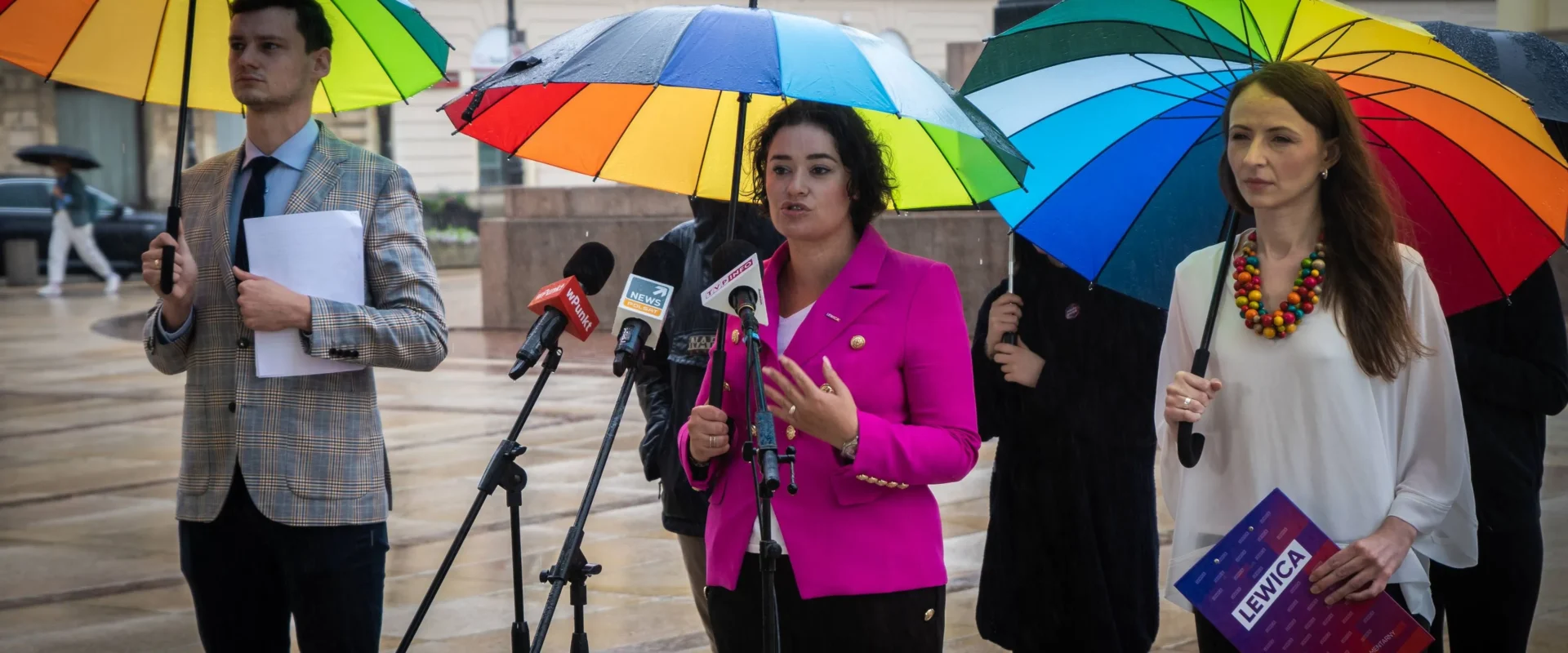 Konferencja prasowa na deszczowym placu, trzy osoby stoją pod kolorowymi parasolkami, a kobieta w różowym żakiecie przemawia do mikrofonów, podczas gdy druga trzyma teczkę z napisem Lewica.