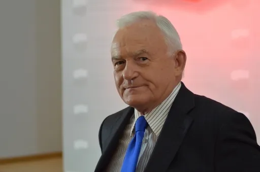 Starszy mężczyzna w ciemnym garniturze i niebieskim krawacie patrzy w stronę aparatu z wyrazem twarzy sugerującym zażenowanie, na jasnym tle.