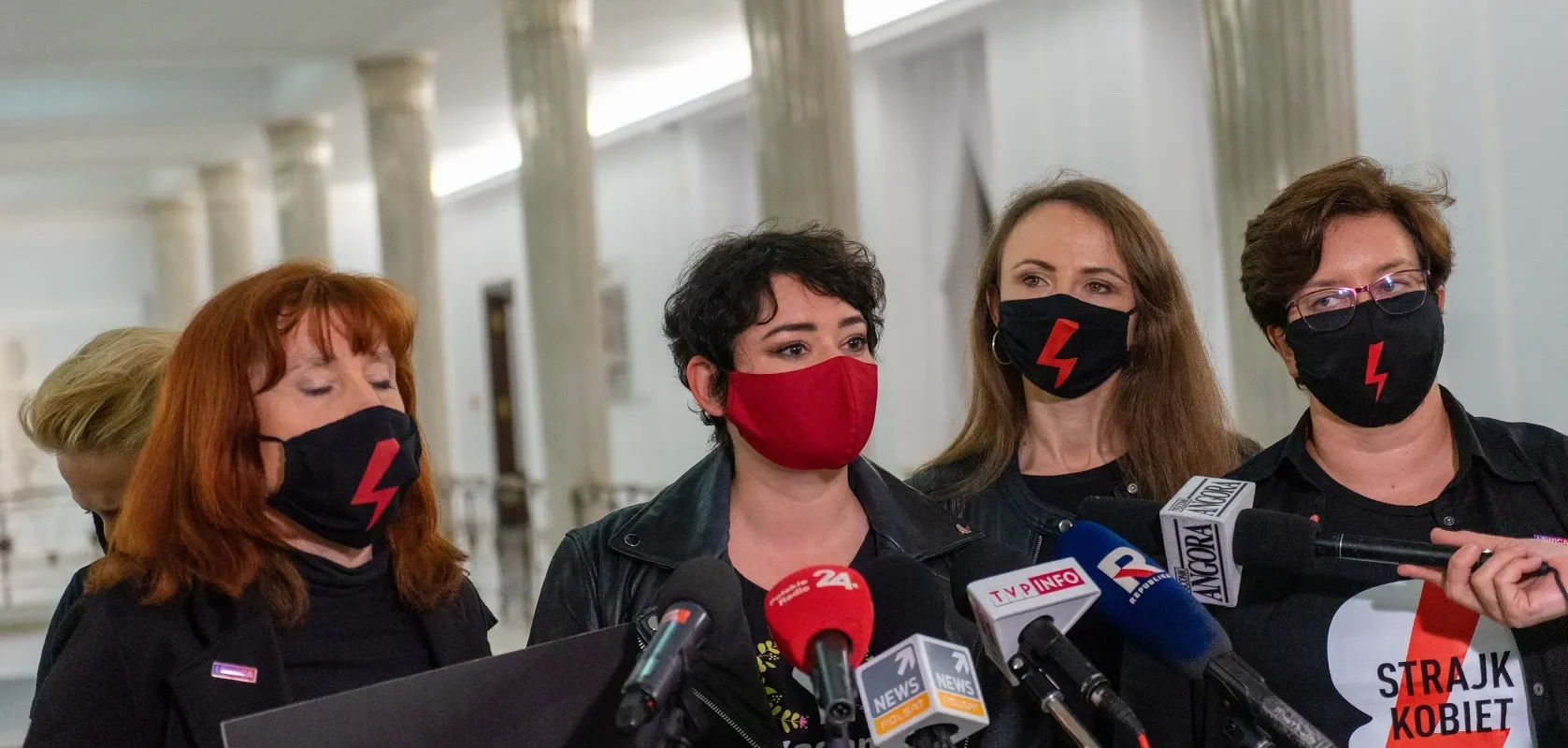Kobiety w czarnych ubraniach i maseczkach z czerwonym błyskawicą stoją przy mikrofonach podczas konferencji prasowej, jedna z nich ma koszulkę z napisem Strajk Kobiet, w tle widoczne kolumny.