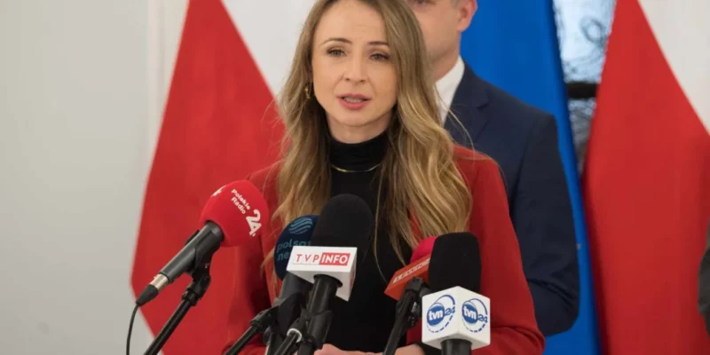 Kobieta w czerwonym żakiecie przemawia na konferencji prasowej, stojąc za zestawem mikrofonów z logotypami mediów, na tle flag państwowych.