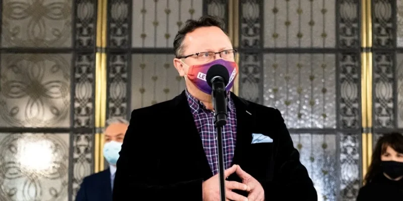 Mężczyzna w czarnej marynarce i koszuli w kratę stoi przy mikrofonie, nosi maseczkę, a w tle widoczne są ozdobne drzwi oraz dwie osoby w maseczkach.