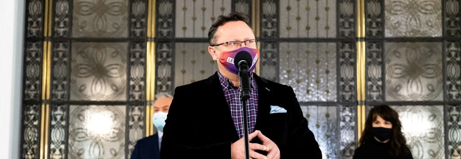 Mężczyzna w czarnej marynarce i koszuli w kratę stoi przy mikrofonie, nosi maseczkę, a w tle widoczne są ozdobne drzwi oraz dwie osoby w maseczkach.