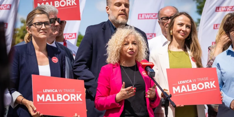 Grupa osób Lewicy na konferencji prasowej w Malborku, na pierwszym planie kobieta w różowym żakiecie przemawia do mikrofonu, wokół plakaty i flagi z logo Lewica.