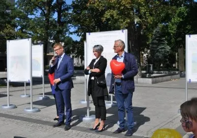 Trzy osoby stoją na miejskim placu w Częstochowie podczas happeningu „SOS – Szukamy Rodzin Zastępczych”: jeden przemawia do mikrofonu, inny trzyma czerwone serce.