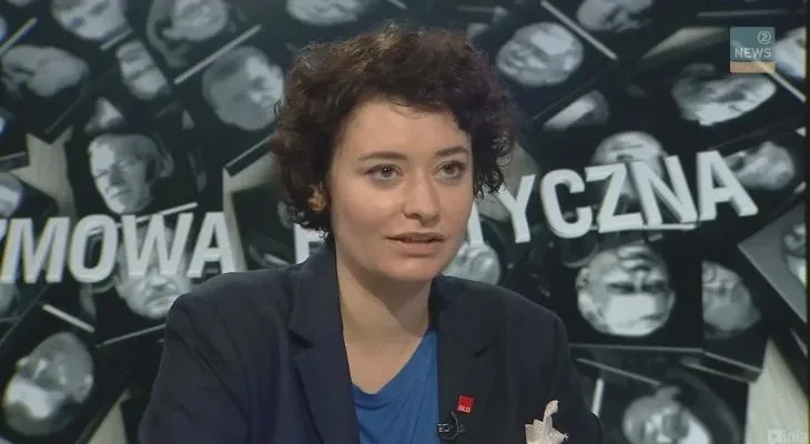 Kobieta w marynarce z krótkimi ciemnymi włosami udziela komentarza w studiu telewizyjnym na tle ekranu z napisami politycznymi, w programie o pozyskiwaniu środków z UE.