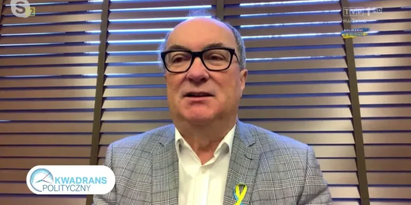 Na zdjęciu widoczny jest mężczyzna w okularach, ubrany w szary wzorzysty garnitur oraz białą koszulę, siedzący przed żaluzjami, który występuje w programie telewizyjnym "Kwadrans Polityczny".