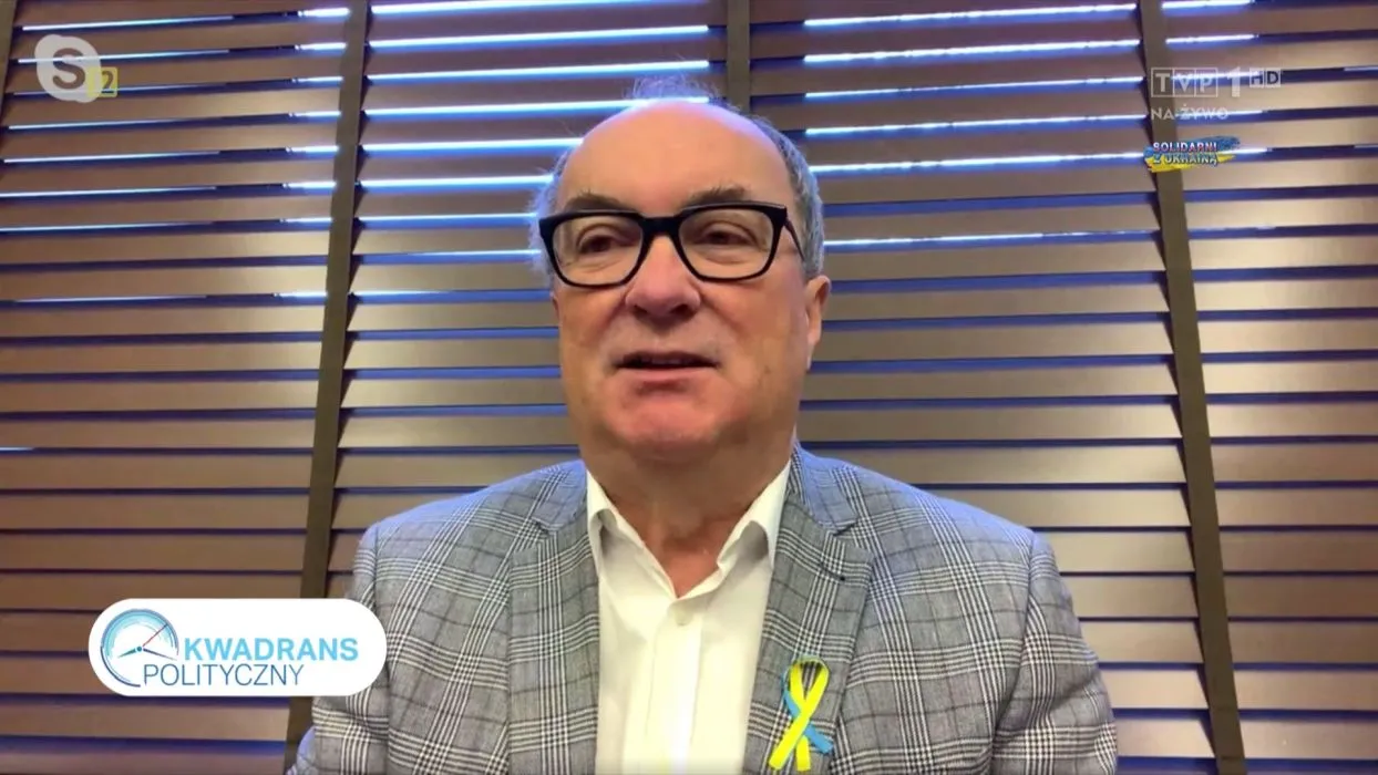 Na zdjęciu widoczny jest mężczyzna w okularach, ubrany w szary wzorzysty garnitur oraz białą koszulę, siedzący przed żaluzjami, który występuje w programie telewizyjnym "Kwadrans Polityczny".