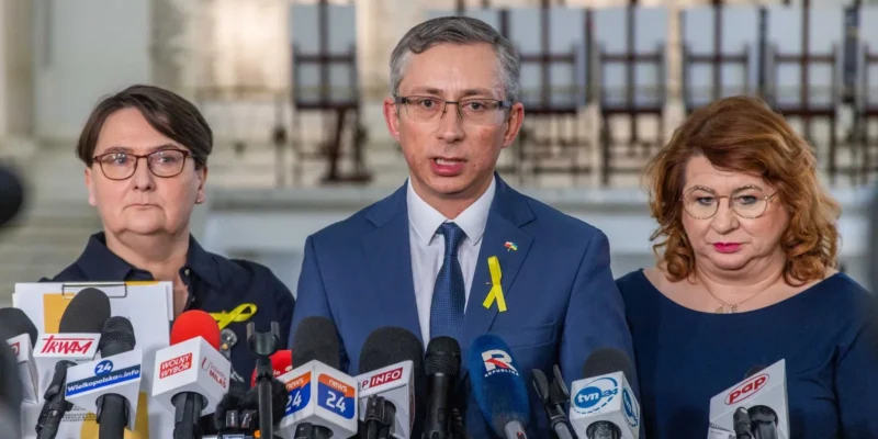 Na zdjęciu przedstawiono trzech mówców stojących przed mikrofonami, z jednym z nich w garniturze, prowadzącego konferencję prasową, podczas gdy dwaj pozostali słuchają.