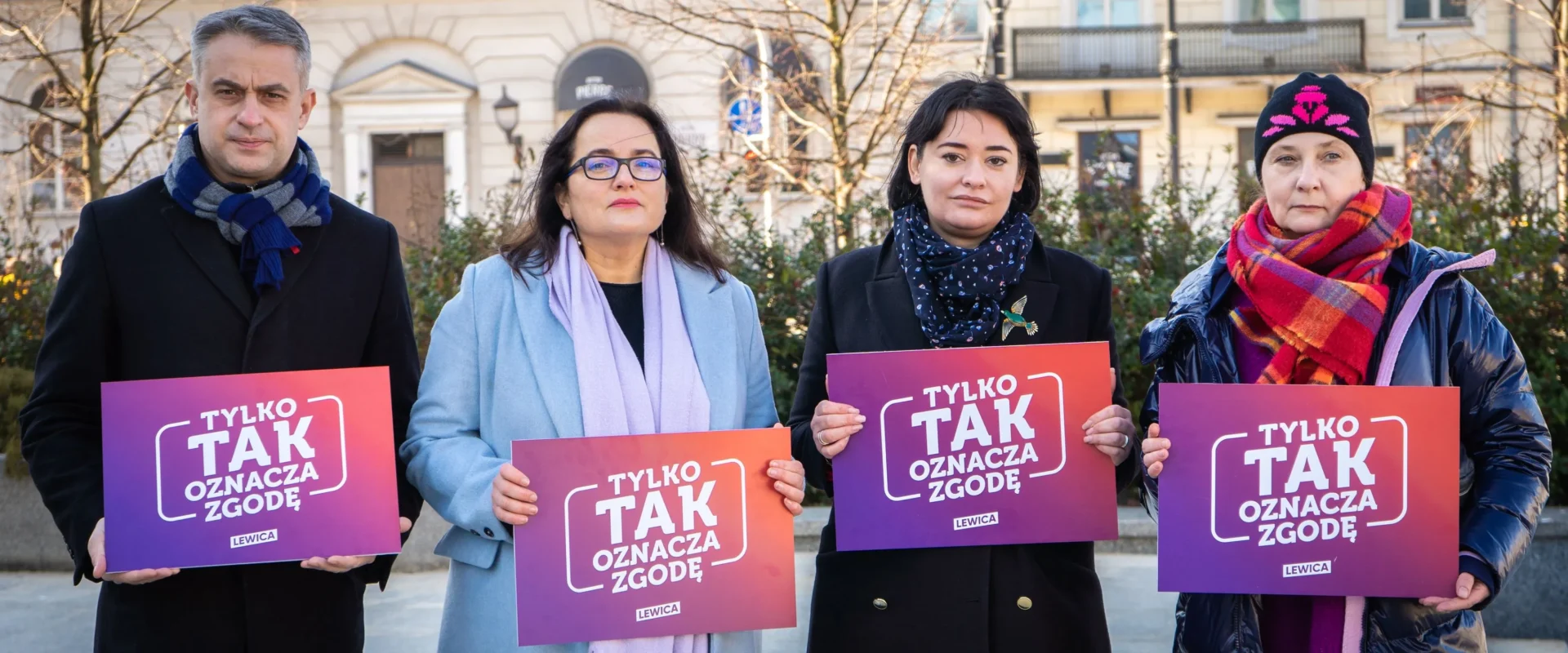 Cztery osoby stoją na placu w mieście, trzymając plakaty z napisem „TYLKO TAK OZNACZA ZGODĘ” w gradientowych kolorach fioletu i pomarańczy.