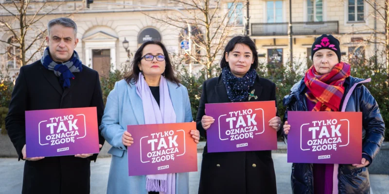 Cztery osoby stoją na placu w mieście, trzymając plakaty z napisem „TYLKO TAK OZNACZA ZGODĘ” w gradientowych kolorach fioletu i pomarańczy.