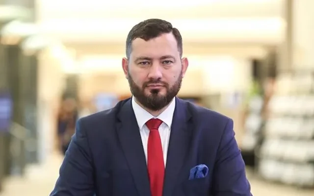 Mężczyzna w granatowym garniturze z czerwonym krawatem i niebieską poszetką stoi na tle jasnego, rozmytego wnętrza.