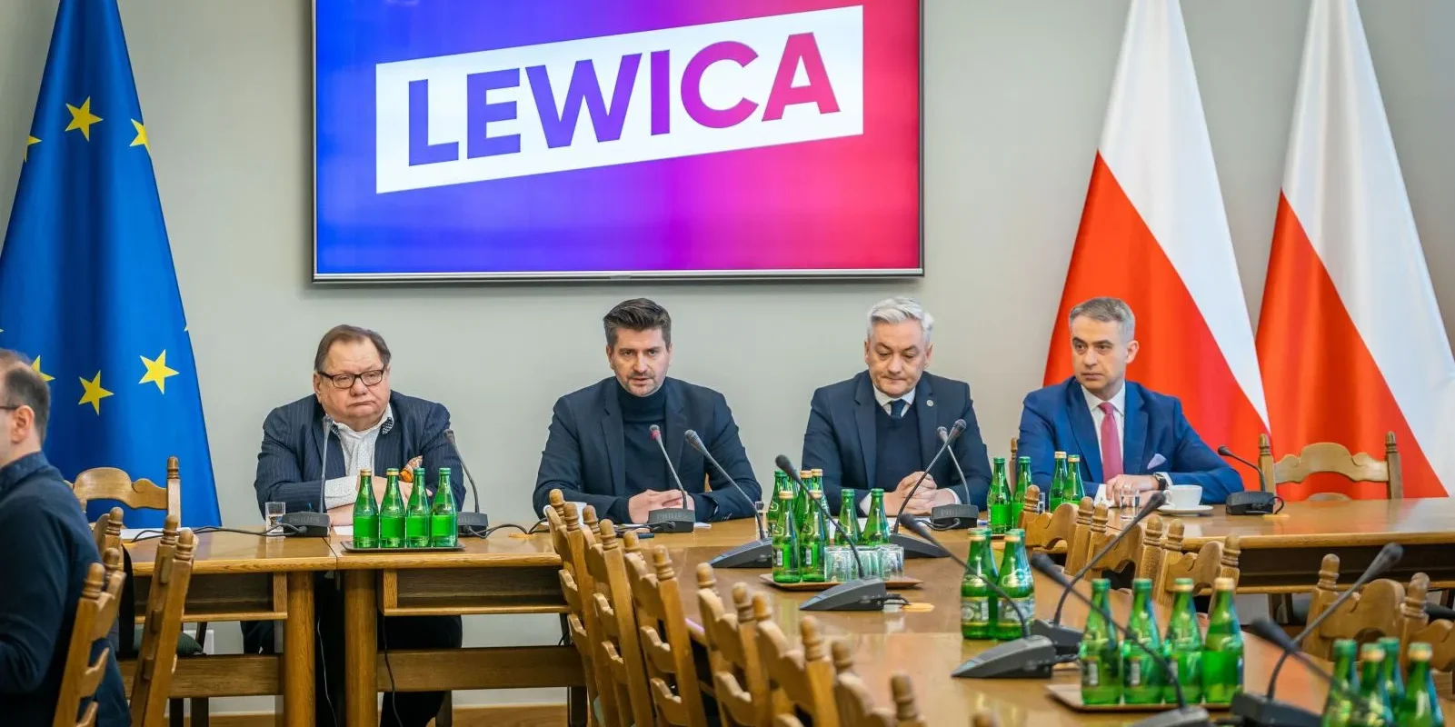 Grupa czterech mężczyzn siedzi przy długim stole podczas konferencji; na ekranie za nimi widnieje napis LEWICA, a po lewej stronie stoją flagi Unii Europejskiej, a po prawej flagi Polski.
