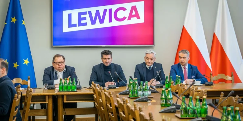 Grupa czterech mężczyzn siedzi przy długim stole podczas konferencji; na ekranie za nimi widnieje napis LEWICA, a po lewej stronie stoją flagi Unii Europejskiej, a po prawej flagi Polski.