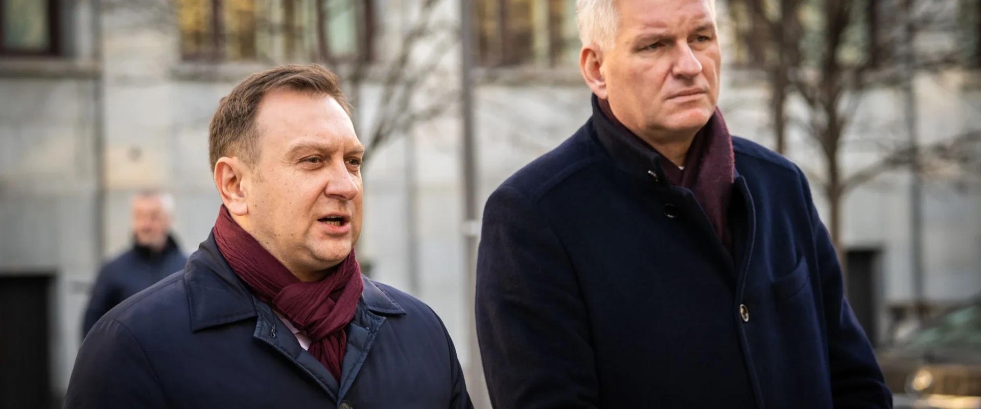 Na zdjęciu widać dwóch mężczyzn w eleganckich ubraniach, stojących na tle budynku, podczas konferencji prasowej.