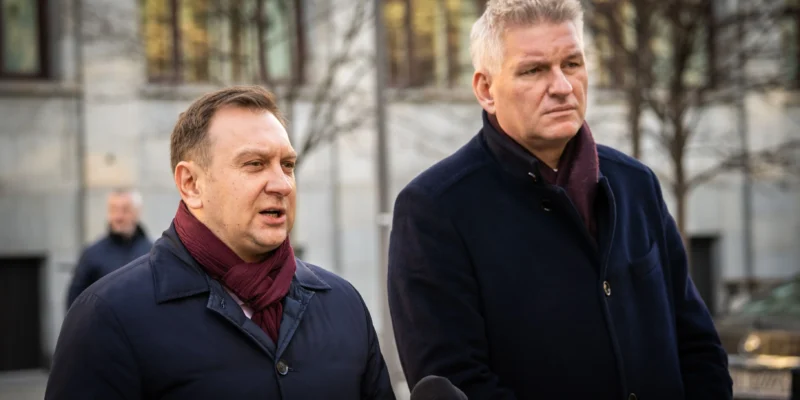 Na zdjęciu widać dwóch mężczyzn w eleganckich ubraniach, stojących na tle budynku, podczas konferencji prasowej.