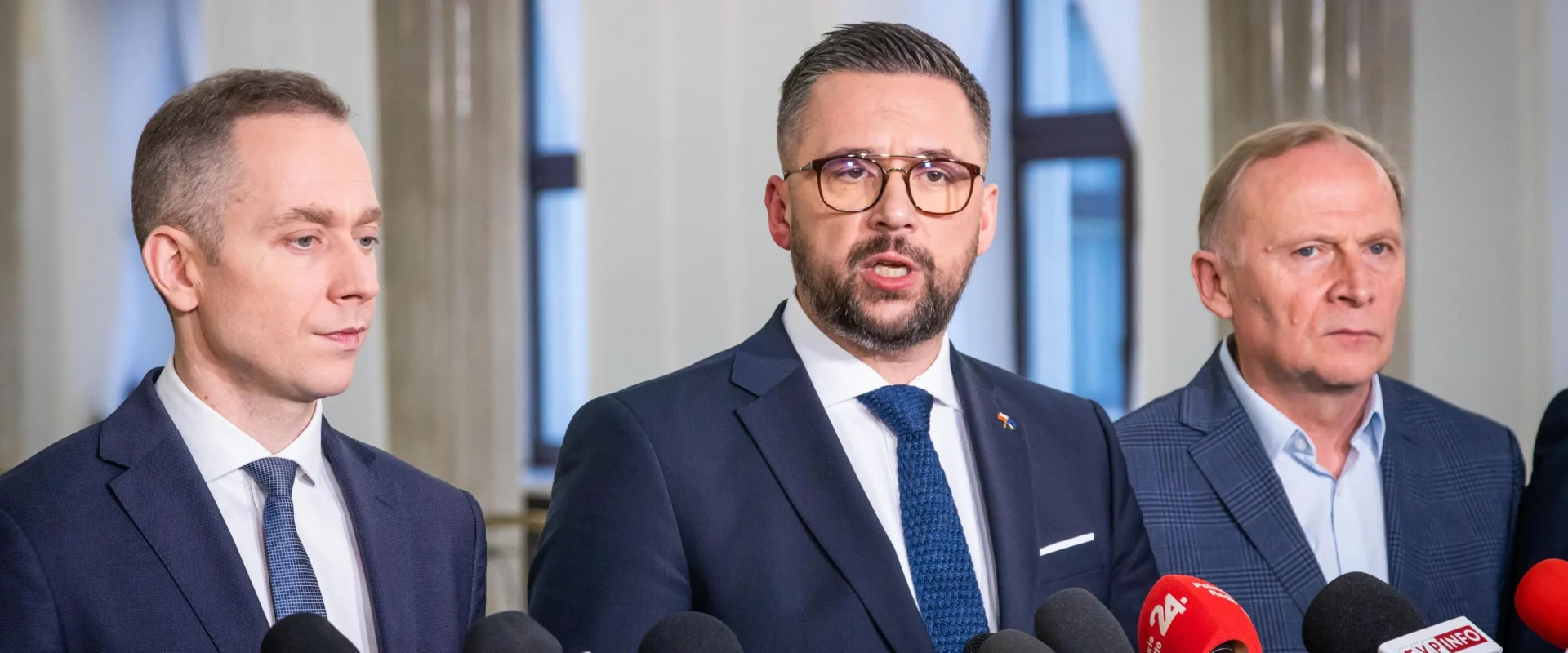 Trzech mężczyzn w garniturach podczas konferencji prasowej o polityce, stojących przed licznymi mikrofonami dziennikarzy.