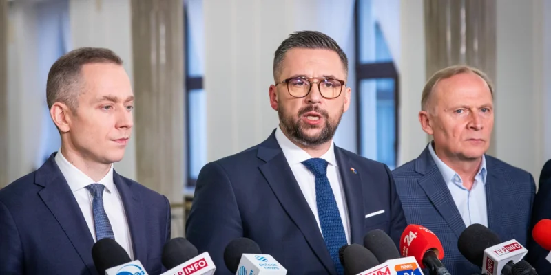 Trzech mężczyzn w garniturach podczas konferencji prasowej o polityce, stojących przed licznymi mikrofonami dziennikarzy.