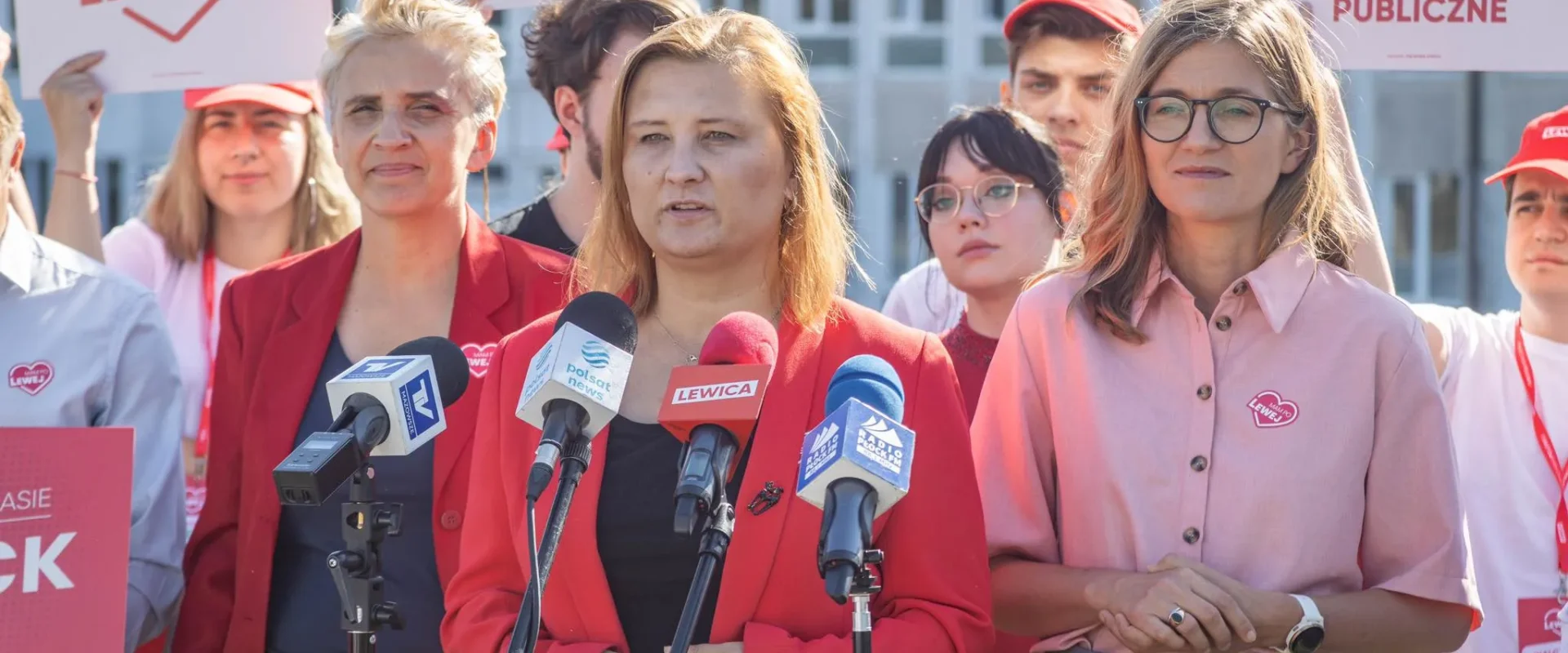 Kobiety działaczki Lewicy przemawiają na konferencji prasowej przed mikrofonom, otoczone plakatami z hasłami i symbolem ruchu Lewica.