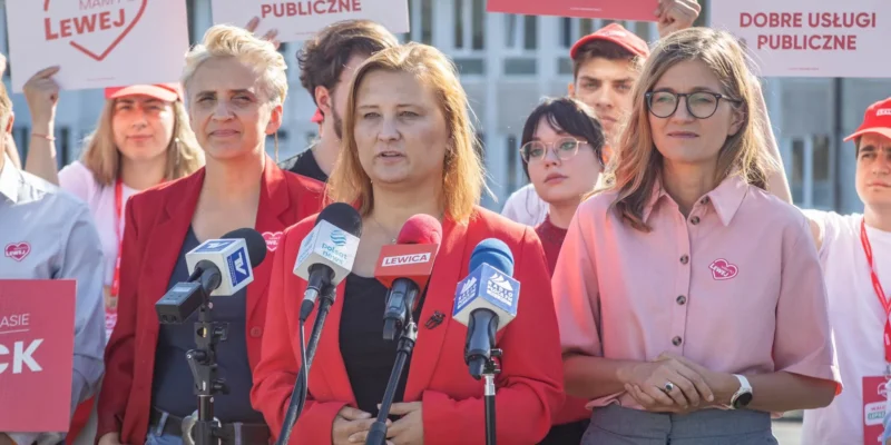 Kobiety działaczki Lewicy przemawiają na konferencji prasowej przed mikrofonom, otoczone plakatami z hasłami i symbolem ruchu Lewica.