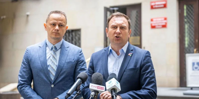 Dwóch mężczyzn w garniturach stoi przed budynkiem, zwracając się do mediów, z mikrofonami przed sobą.