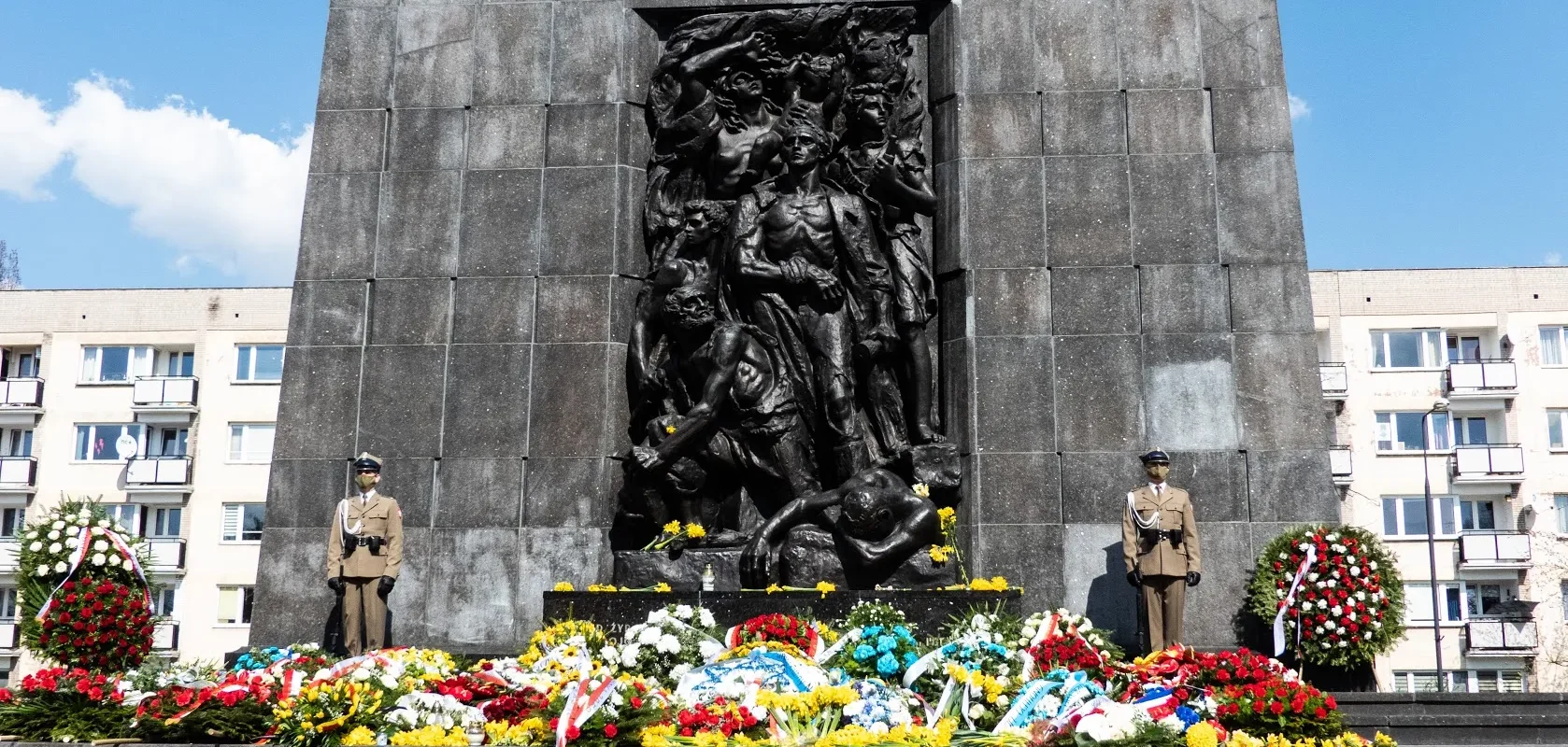 Monument upamiętniający 78. rocznicę Powstania w Getcie Warszawskim z ciemnym reliefem przedstawiającym grupę postaci, otoczony kolorowymi kwiatami i wieńcami; po obu stronach stoją żołnierze w mundurach, a w tle widać bloki mieszkalne.