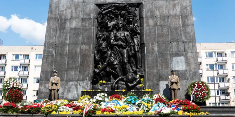 Monument upamiętniający 78. rocznicę Powstania w Getcie Warszawskim z ciemnym reliefem przedstawiającym grupę postaci, otoczony kolorowymi kwiatami i wieńcami; po obu stronach stoją żołnierze w mundurach, a w tle widać bloki mieszkalne.