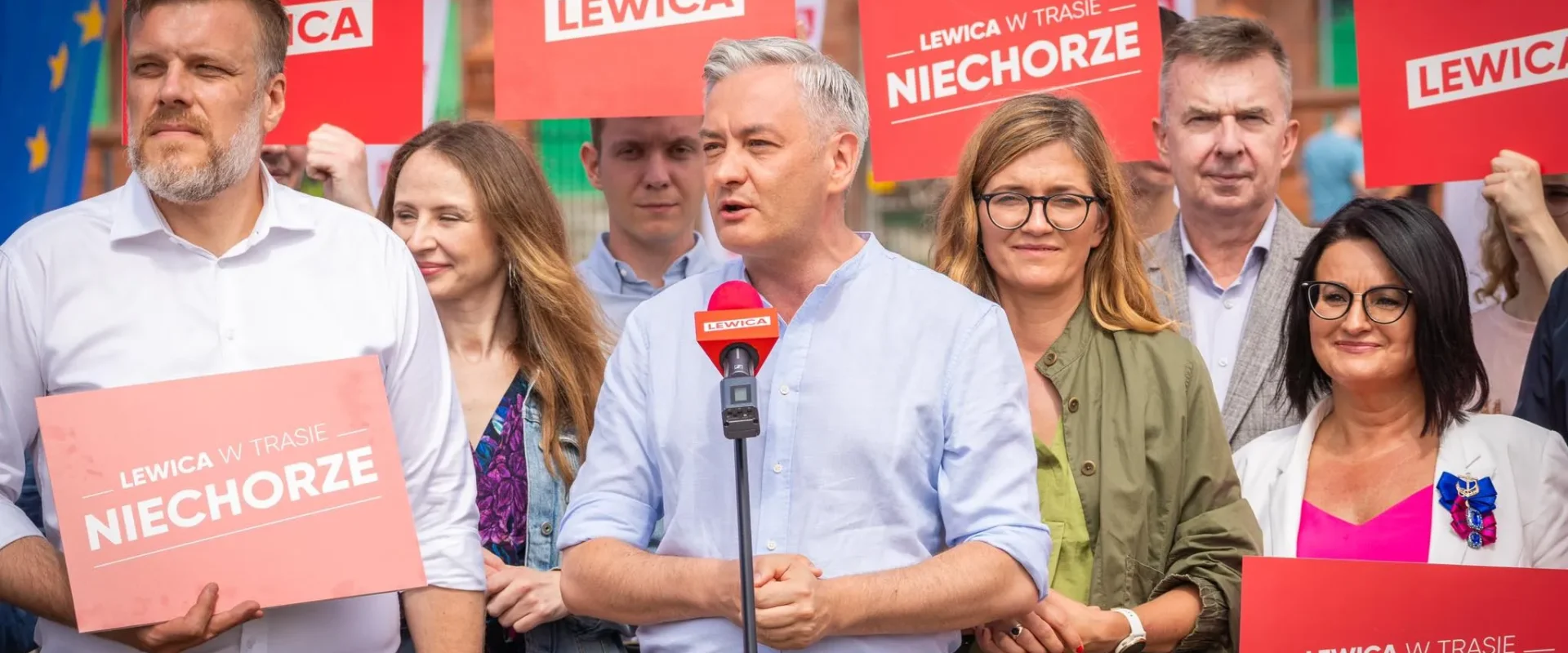 Grupa polityków Lewicy podczas konferencji prasowej; mężczyzna przemawia do mikrofonu, otoczony innymi zwolennikami, na tle czerwonych plakatów z napisem LEWICA.