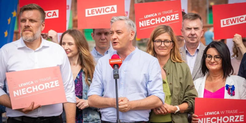 Grupa polityków Lewicy podczas konferencji prasowej; mężczyzna przemawia do mikrofonu, otoczony innymi zwolennikami, na tle czerwonych plakatów z napisem LEWICA.