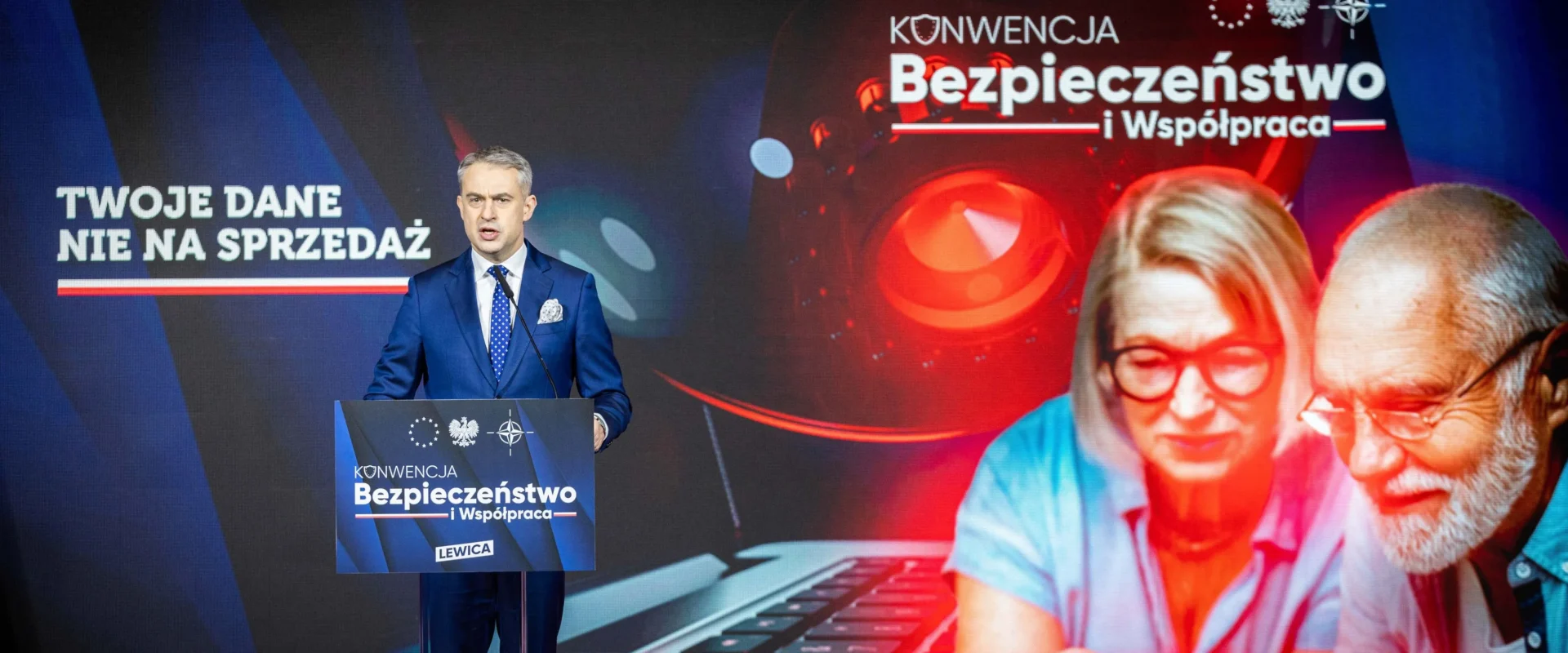 Mężczyzna w granatowym garniturze przemawia z mównicy podczas konferencji „Bezpieczeństwo i Współpraca”, na tle baneru z napisem „Twoje dane nie na sprzedaż” i wizerunkiem starszej pary oglądającej laptopa.