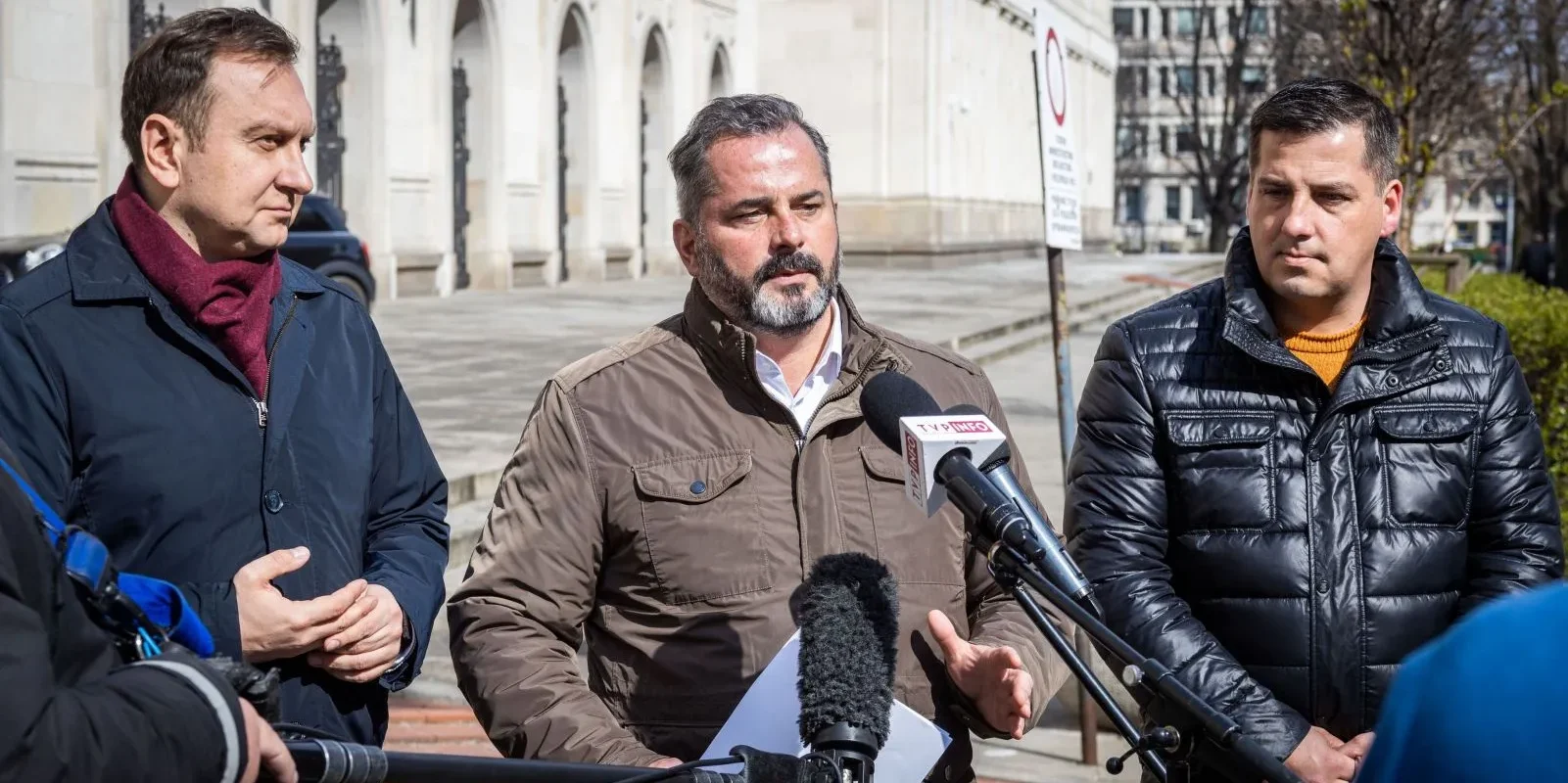Trzech mężczyzn w kurtkach stoi na zewnątrz budynku i udziela konferencji prasowej, mówiąc do mikrofonów.