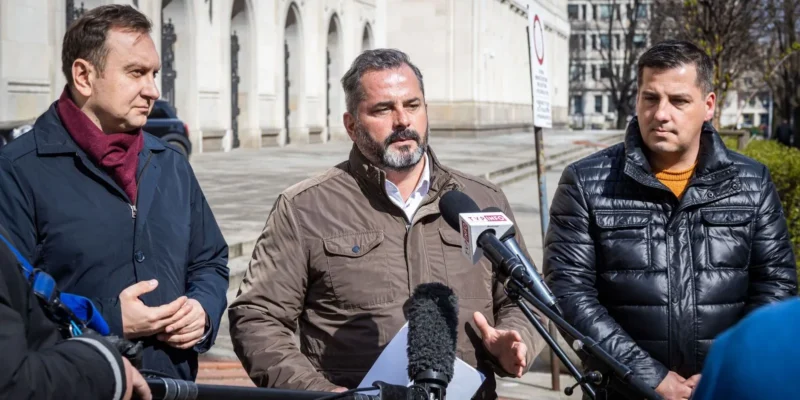 Trzech mężczyzn w kurtkach stoi na zewnątrz budynku i udziela konferencji prasowej, mówiąc do mikrofonów.